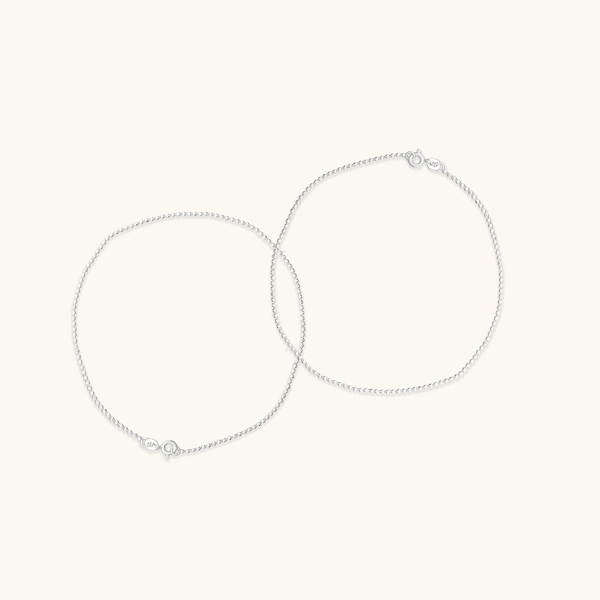 Two fine, shimmering silver chain anklet  positioned in overlapping circles to demonstrate their delicate texture and clasp hardware.