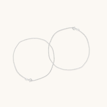 Two fine, shimmering silver chain anklet  positioned in overlapping circles to demonstrate their delicate texture and clasp hardware.