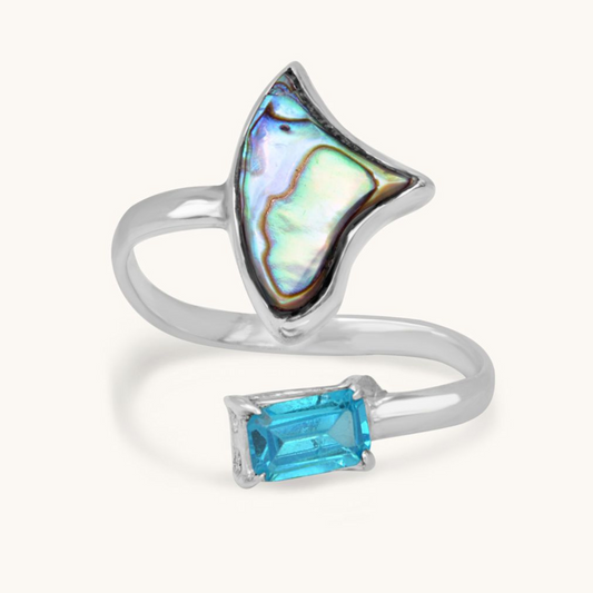 A front-facing studio shot of an adjustable open silver ring, showcasing a large triangular iridescent abalone shell inlay at one end and a rectangular-cut vibrant blue gemstone at the other.