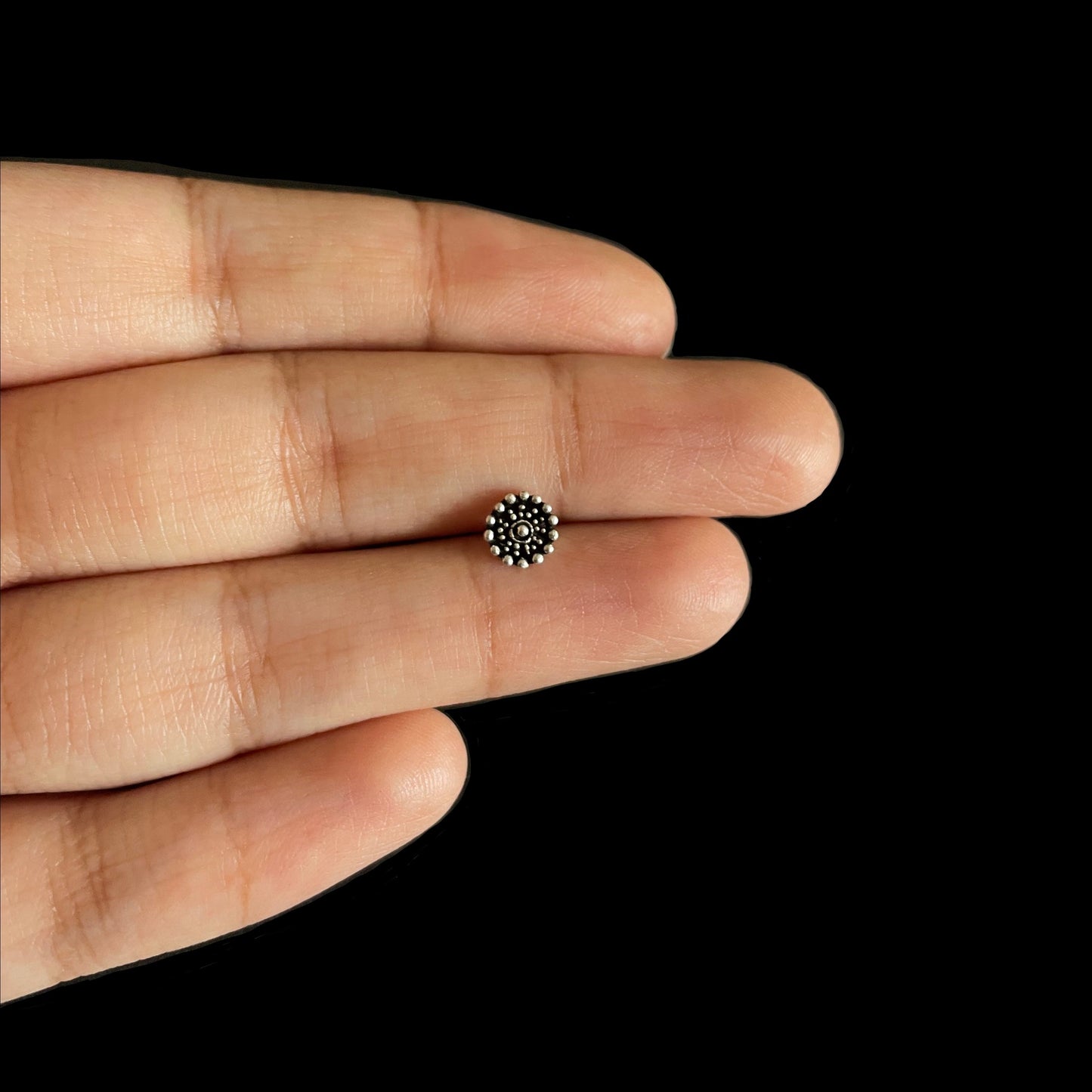 A close-up view of a small, intricate circular silver nose pin featuring a decorative beaded pattern, held between two fingers.