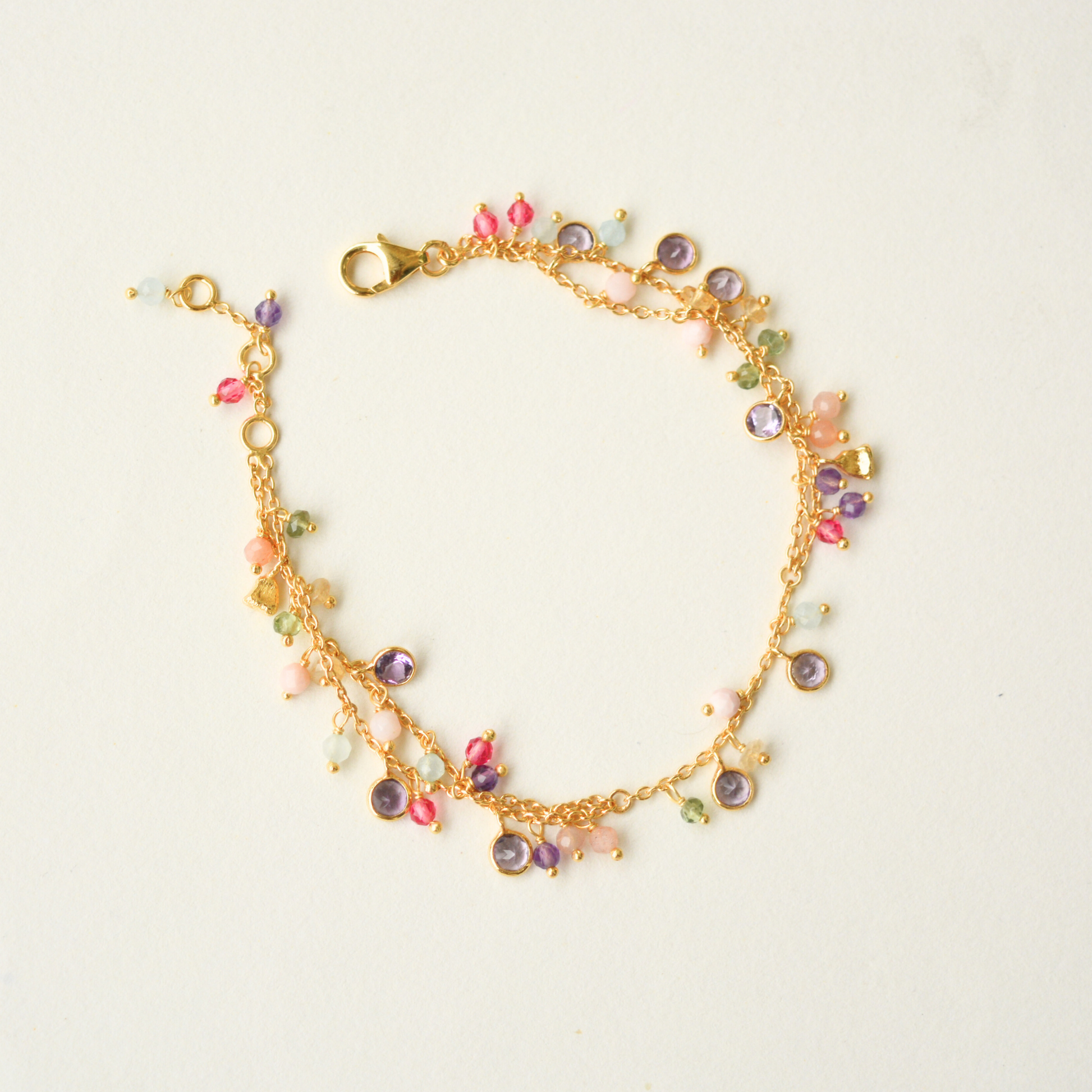 A delicate gold-plated silver double-chain bracelet adorned with an assortment of small, colorful gemstone charms, set against a white background.