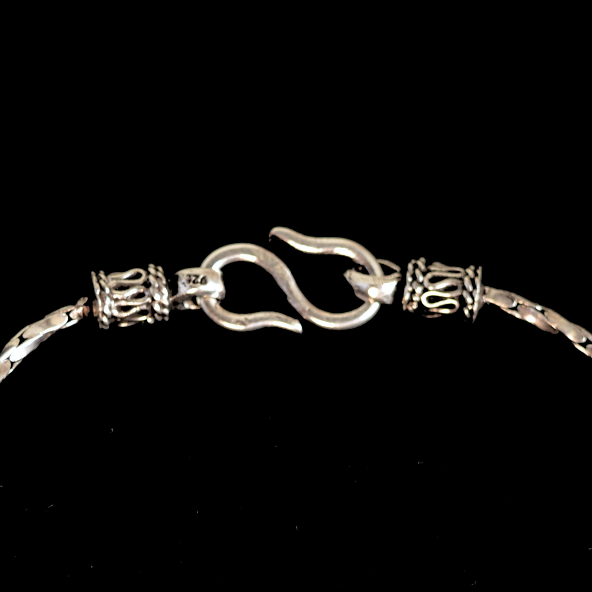 A macro detail shot showing the S-hook clasp and intricate metalwork ends of a polished silver link bracelet against a black backdrop.