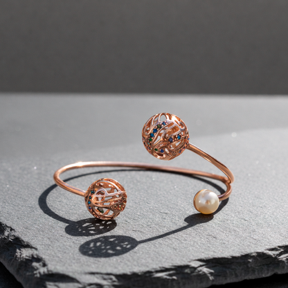 Rose gold Rolling Cuff Bracelet featuring open globe detailing with colorful accents and a pearl end, styled on a stone slab background.