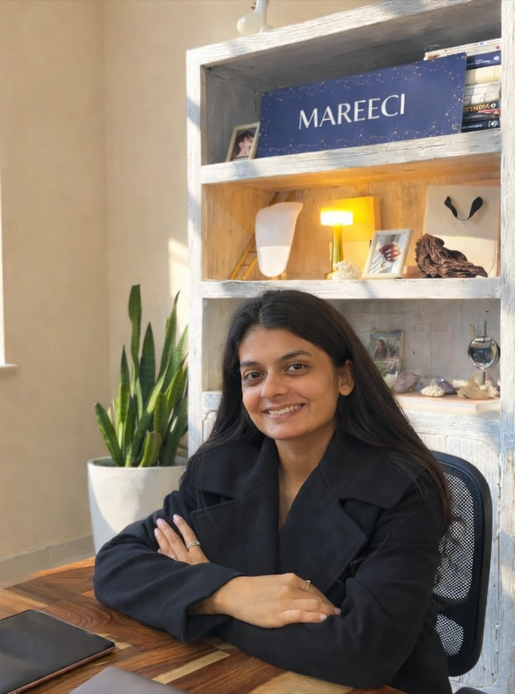 A woman with long dark hair sits at a wooden desk in her DESIGN STUDIO, wearing a black blazer. Behind her is a white shelving unit with a blue sign reading 'MAREECI' 