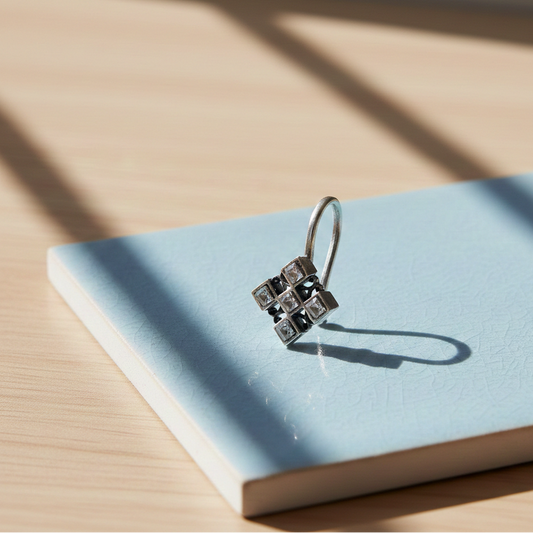 Silver Mirage Clip-On nose jewelry featuring a square geometric design with inset crystals, displayed on a soft blue ceramic tile in natural sunlight.