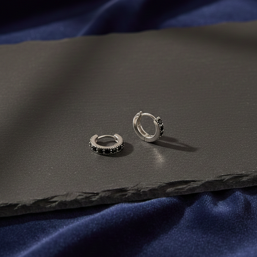Silver huggie hoop earrings with black stones, displayed on a dark textured surface with blue fabric background.