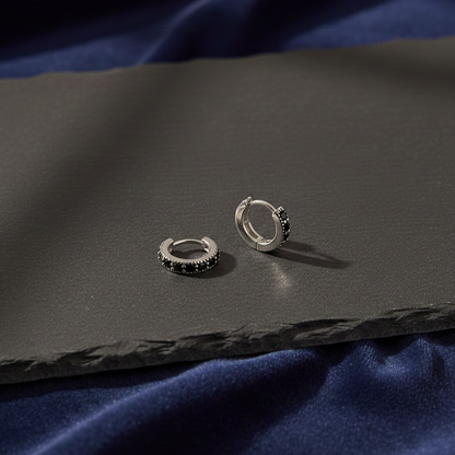 Silver huggie hoop earrings with black stones, displayed on a dark textured surface with blue fabric background.