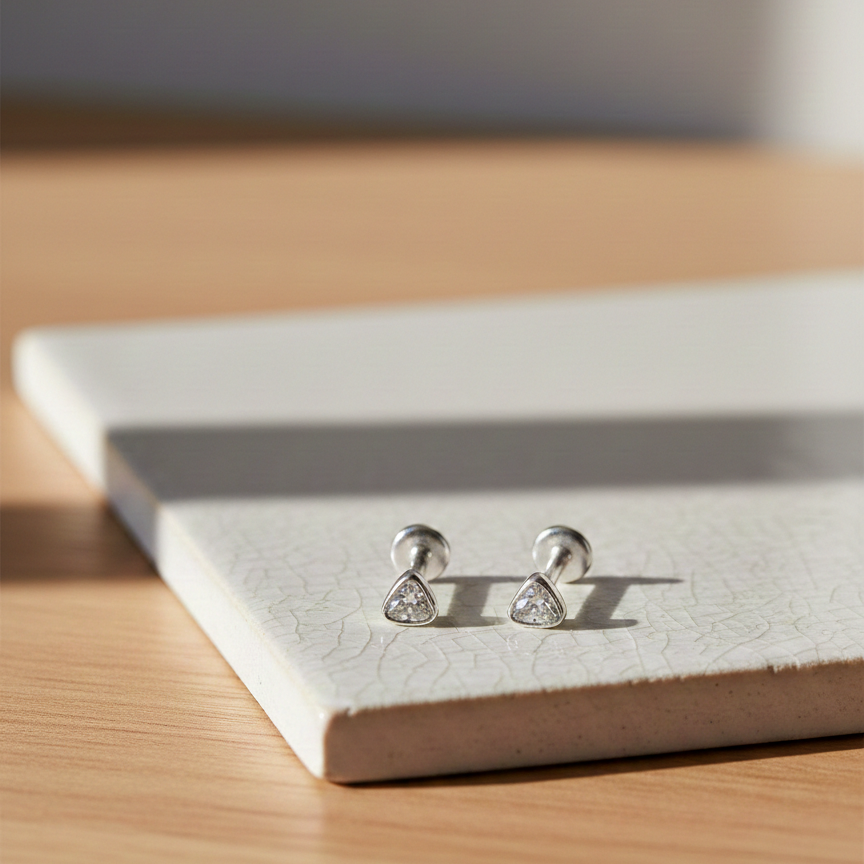 Pair of silver Trillion Flatback Earrings featuring small triangular gemstone studs, displayed on a textured ceramic tile in warm natural light.