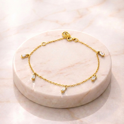 Dainty yellow gold Floating Stone Bracelet with five suspended baguette diamond-style charms, shown resting on a white marble display stand with a soft sunlit background.