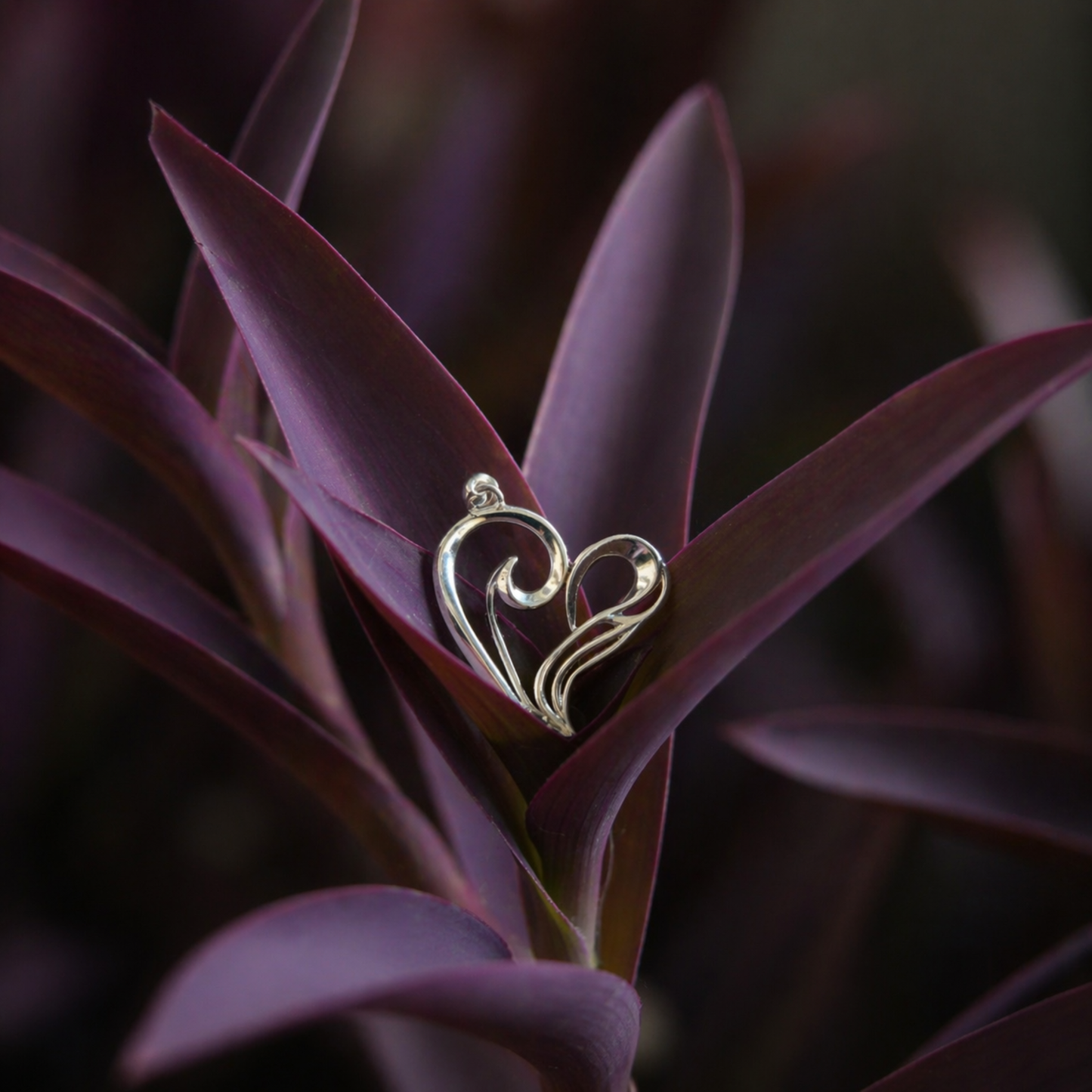 A handcrafted 925 sterling silver A Little Love Pendant with a modern open wired heart design, nestled within deep purple leaves for a romantic lifestyle shot.
