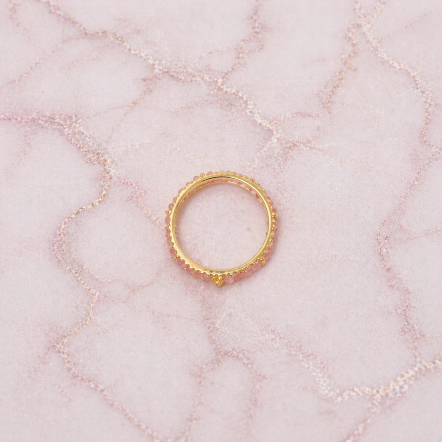 An overhead view of the Blush Ball Ring, showcasing a continuous band of tiny pink tourmaline beads set in 18kt gold plated 925 sterling silver on a pink marbled background.