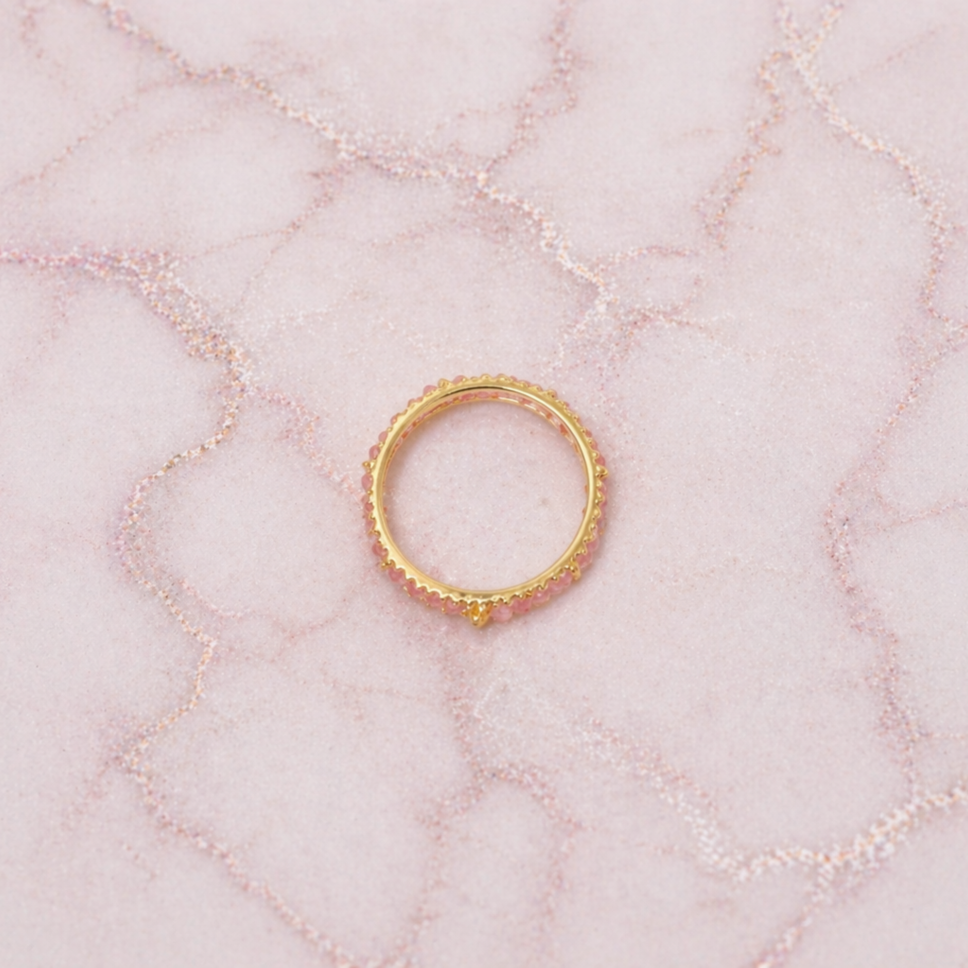 An overhead view of the Blush Ball Ring, showcasing a continuous band of tiny pink tourmaline beads set in 18kt gold plated 925 sterling silver on a pink marbled background.