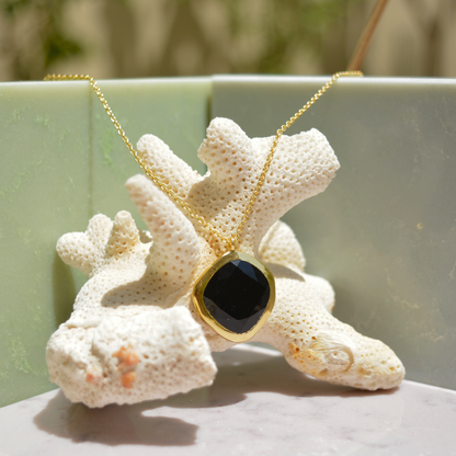 A handcrafted 18kt gold plated Bold Calm necklace featuring an organic square black onyx stone, displayed on a white coral prop against a minimal sage and marble background.