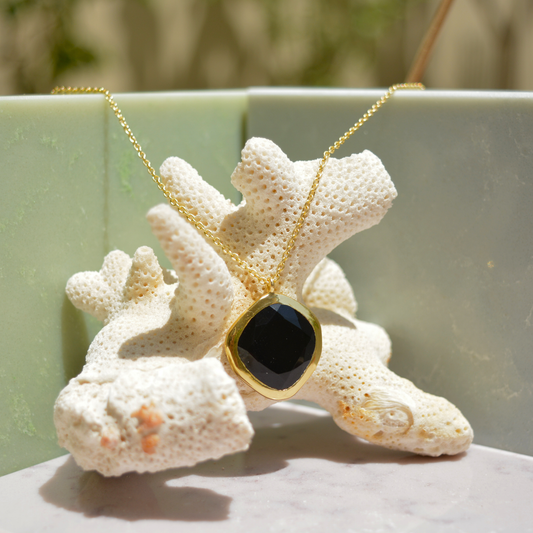A handcrafted 18kt gold plated Bold Calm necklace featuring an organic square black onyx stone, displayed on a white coral prop against a minimal sage and marble background.