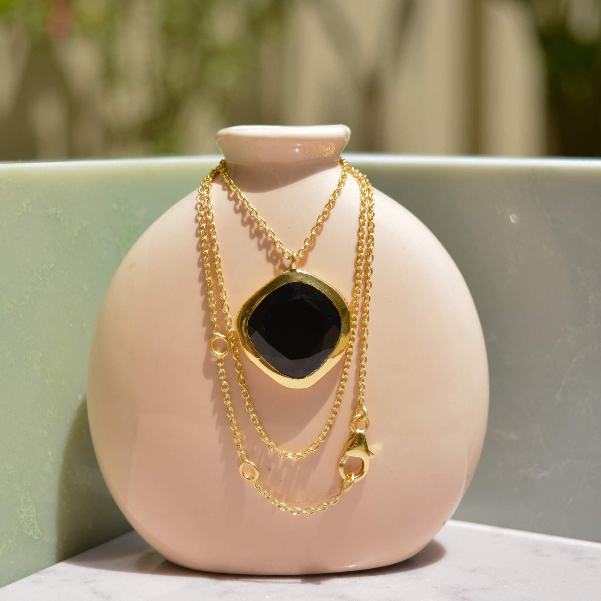 A handcrafted 18kt gold plated Bold Calm necklace with an organic square black onyx stone, draped over a minimal beige ceramic vase.