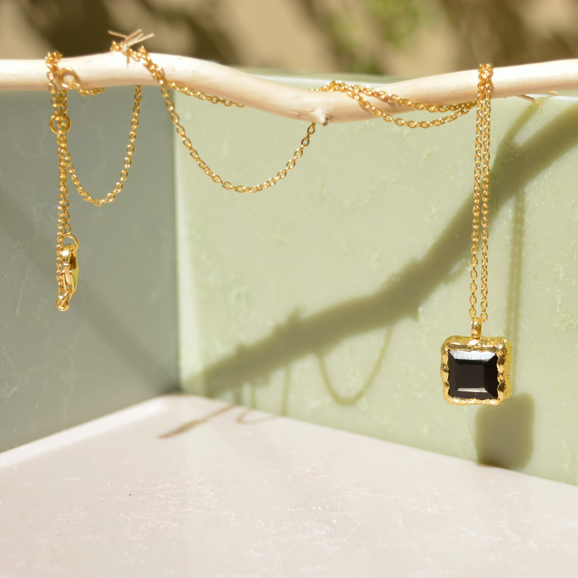 A handcrafted 18kt gold plated Bold Simplicity necklace with a square black onyx pendant, draped over a natural wooden branch against a soft green minimal background.