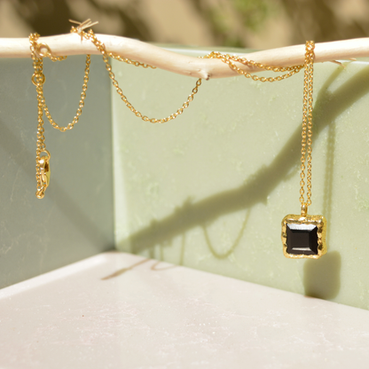 A handcrafted 18kt gold plated Bold Simplicity necklace with a square black onyx pendant, draped over a natural wooden branch against a soft green minimal background.