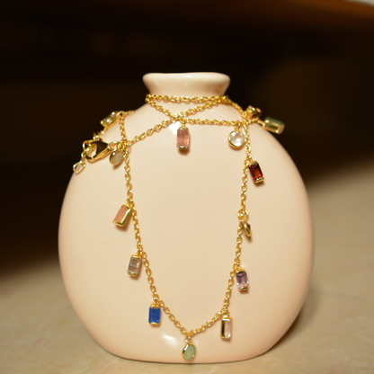 A handcrafted 18kt gold plated A Celebration of Color necklace featuring 13 natural multi-gemstones in rectangular and circular shapes, draped over a minimal beige ceramic vase.