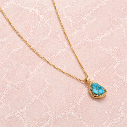 A handcrafted 18kt gold plated Earthy Charm necklace featuring an organic oval copper turquoise stone with natural gold-toned patterns, displayed on a soft pink textured surface.