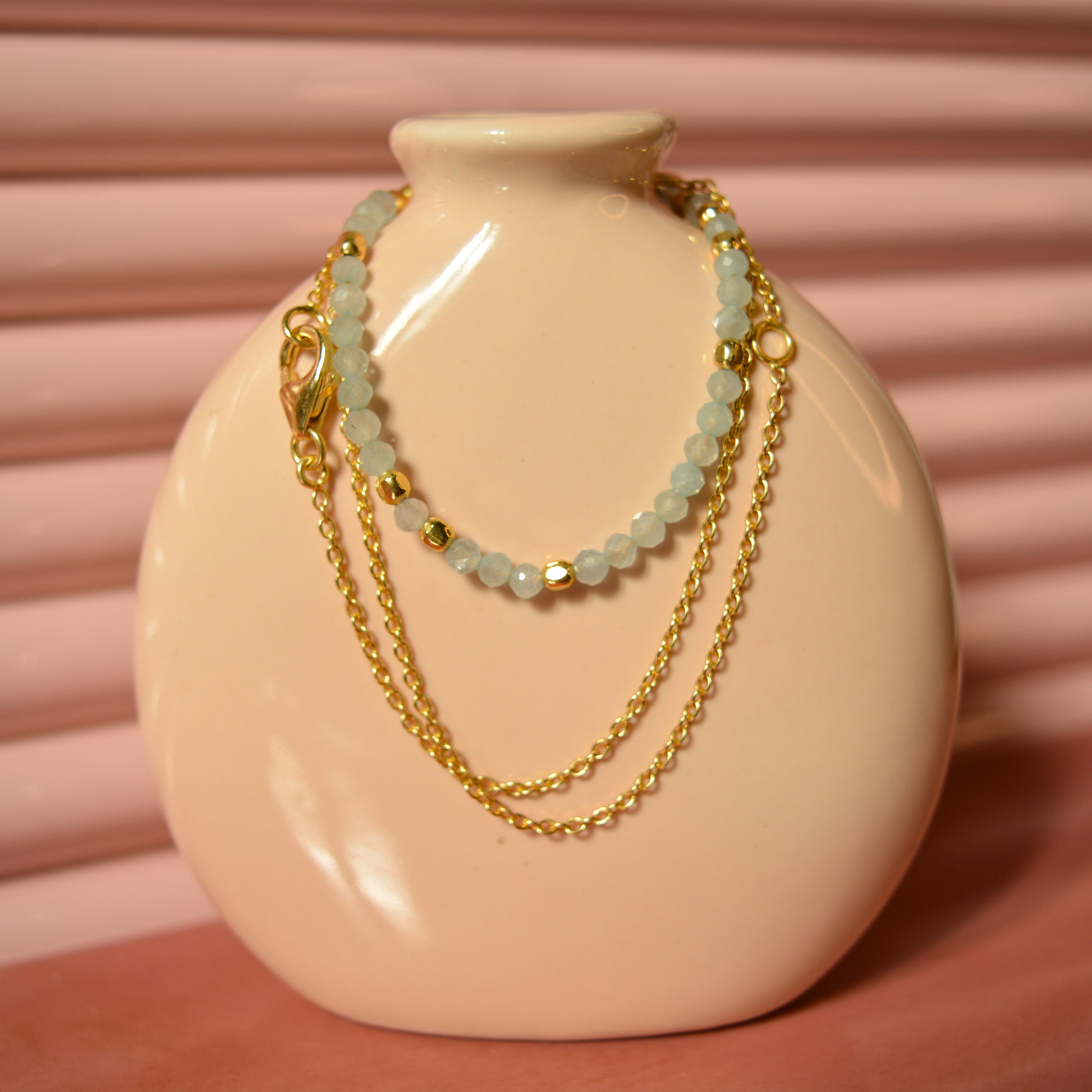 A handcrafted 18kt gold plated Earthy Ease necklace featuring soft blue-grey granite beads, draped over a minimal beige ceramic vase for a contemporary lifestyle aesthetic.
