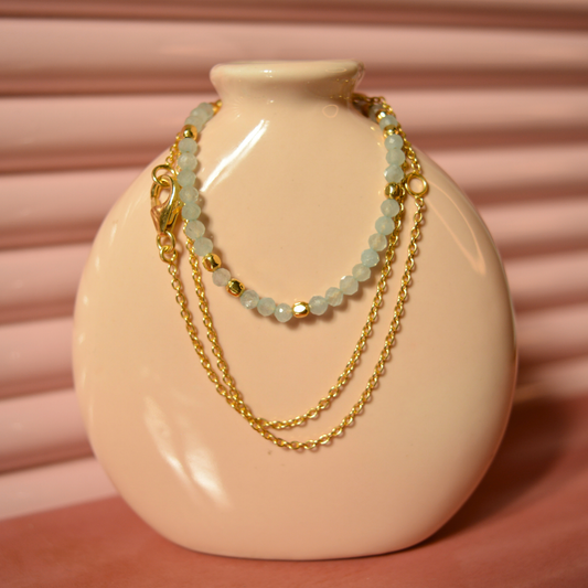 A handcrafted 18kt gold plated Earthy Ease necklace featuring soft blue-grey granite beads, draped over a minimal beige ceramic vase for a contemporary lifestyle aesthetic.