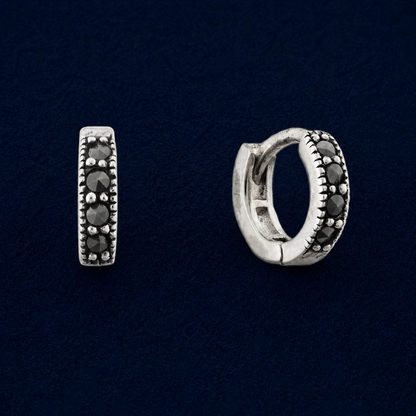 A pair of handcrafted Eclipse mini hoop earrings in 925 sterling silver, featuring an antiqued finish and a row of four round black cubic zirconia stones on a dark navy background.