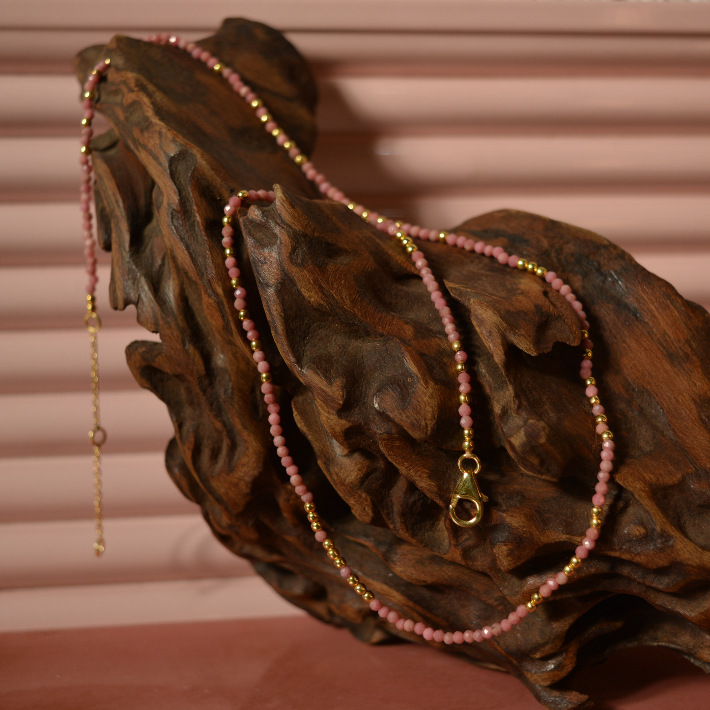 A handcrafted 18kt gold plated Everyday Glow necklace featuring delicate pink multi-stone beads, draped over a natural dark wood sculpture for a rustic lifestyle aesthetic.
