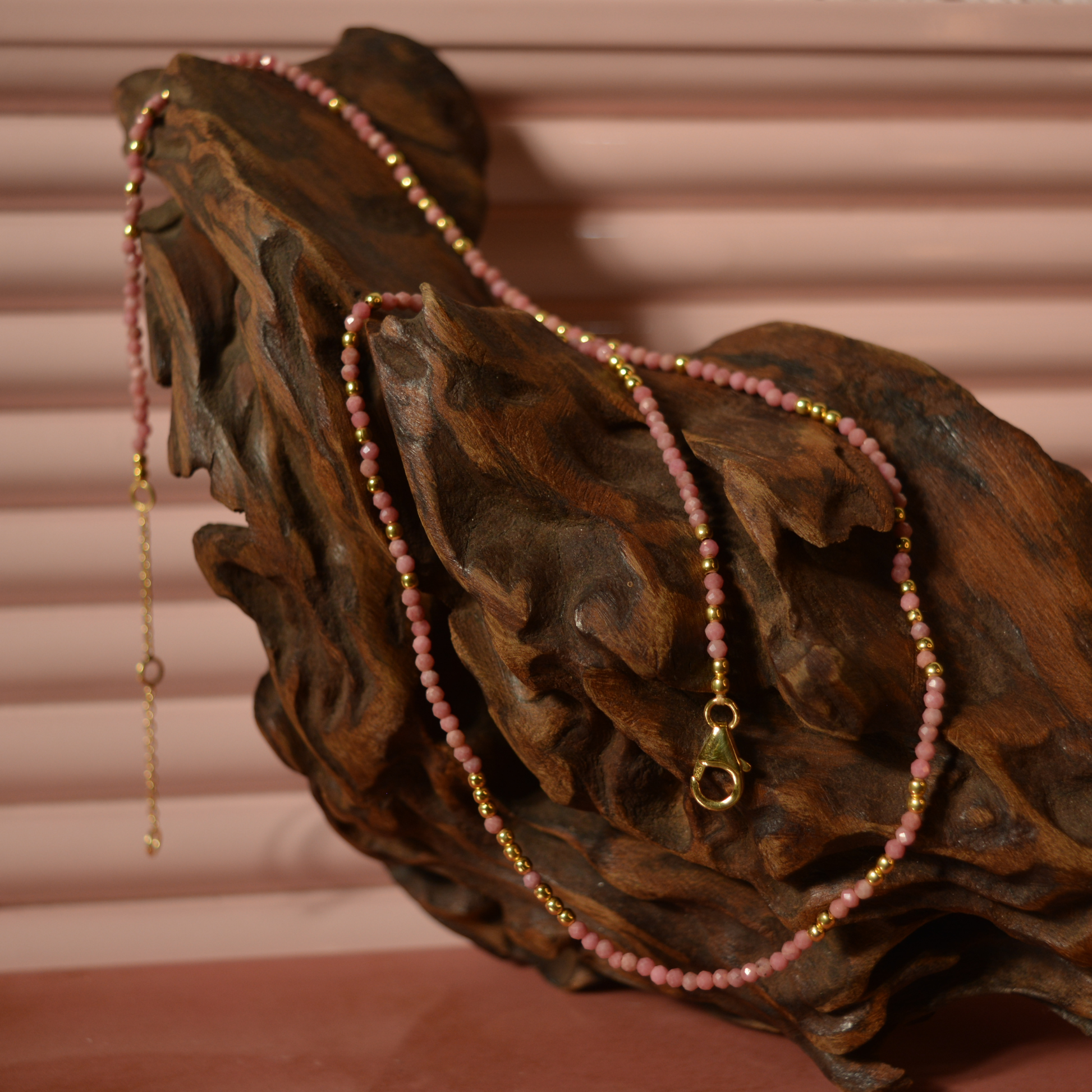 A handcrafted 18kt gold plated Everyday Glow necklace featuring delicate pink multi-stone beads, draped over a natural dark wood sculpture for a rustic lifestyle aesthetic.
