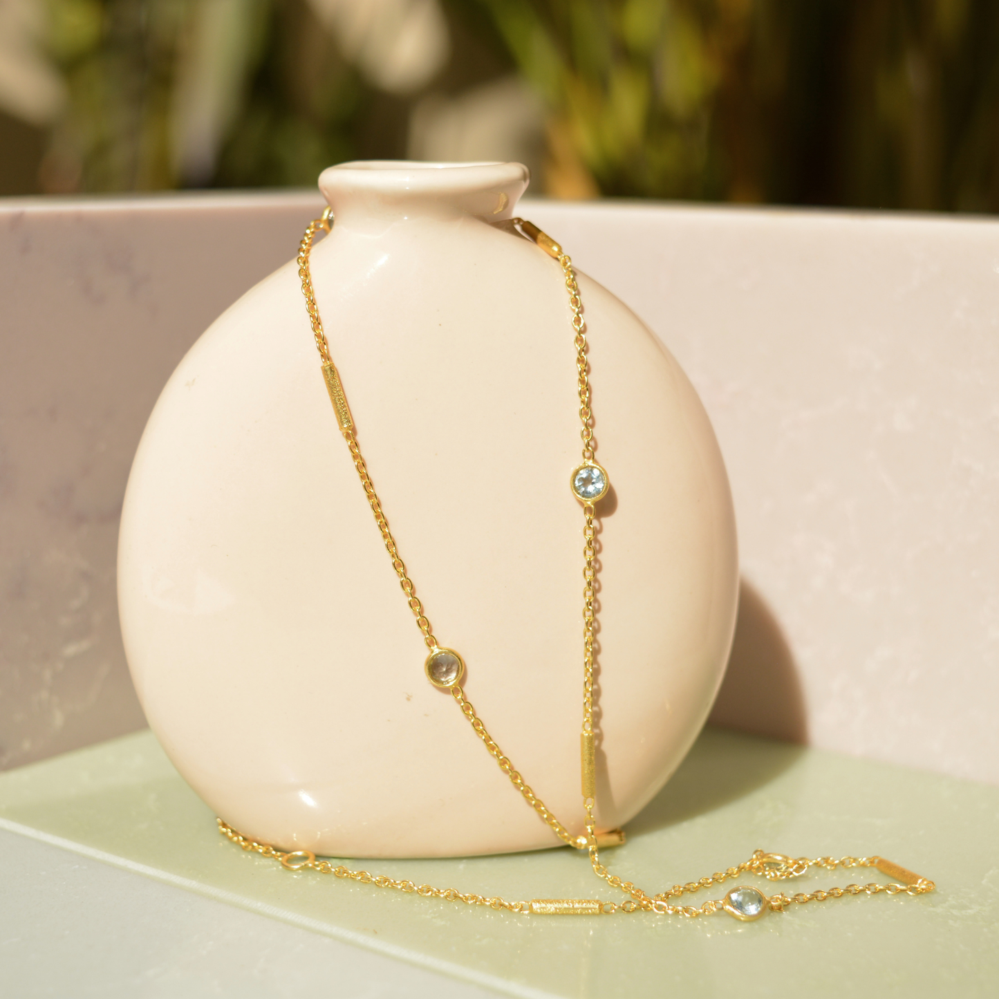 A handcrafted 18kt gold plated Fresh Glow Necklace featuring round blue topaz stones and gold cylindrical bars, draped elegantly over a minimal beige ceramic vase.
