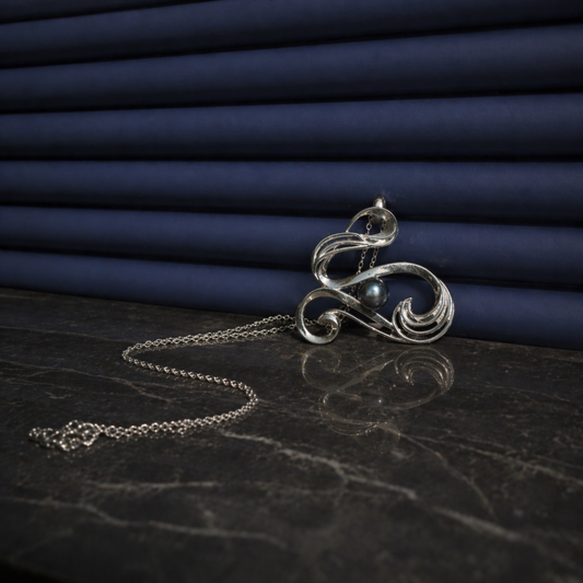 A handcrafted 925 sterling silver Graceful Flow Necklace featuring a wavy pendant with a blue water pearl, displayed on a dark marble surface with a soft reflection and blue textured background.