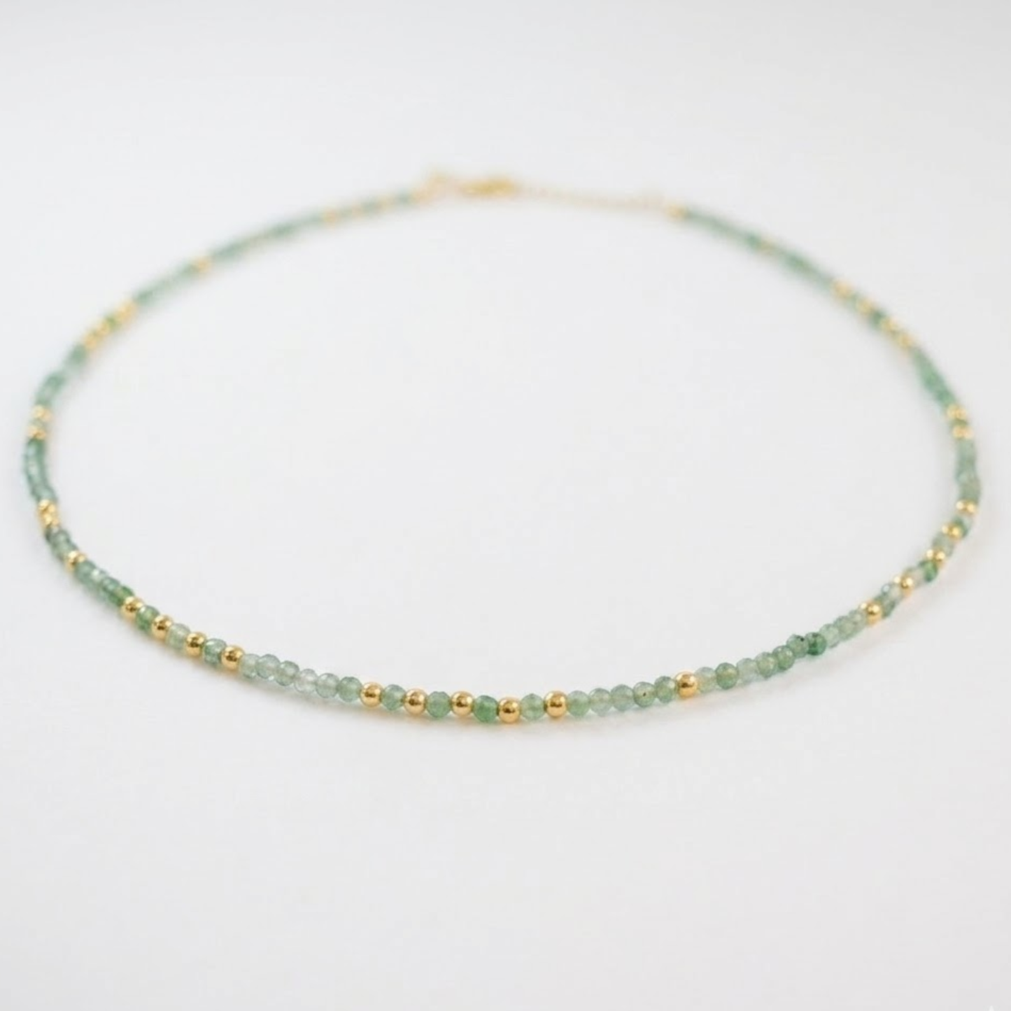 A perspective shot of the Nature’s Calm necklace on a white surface, highlighting the alternating pattern of small faceted green beryl, labradorite, and gold-plated sterling silver beads.