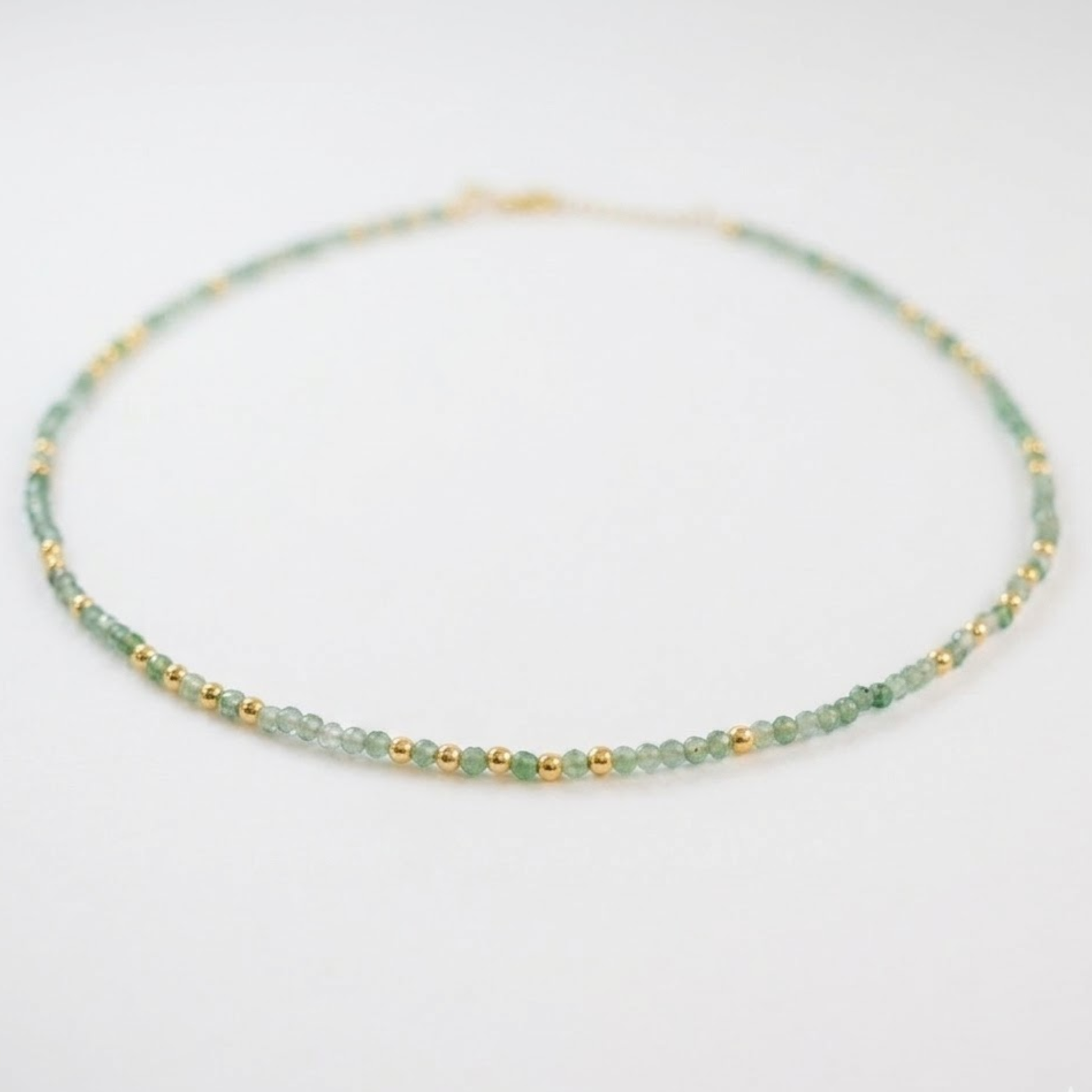 A perspective shot of the Nature’s Calm necklace on a white surface, highlighting the alternating pattern of small faceted green beryl, labradorite, and gold-plated sterling silver beads.