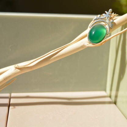 The Natural Elegance green onyx pendant resting on a light wooden branch with soft shadows, highlighting the intricate silver filigree design and rich green stone.