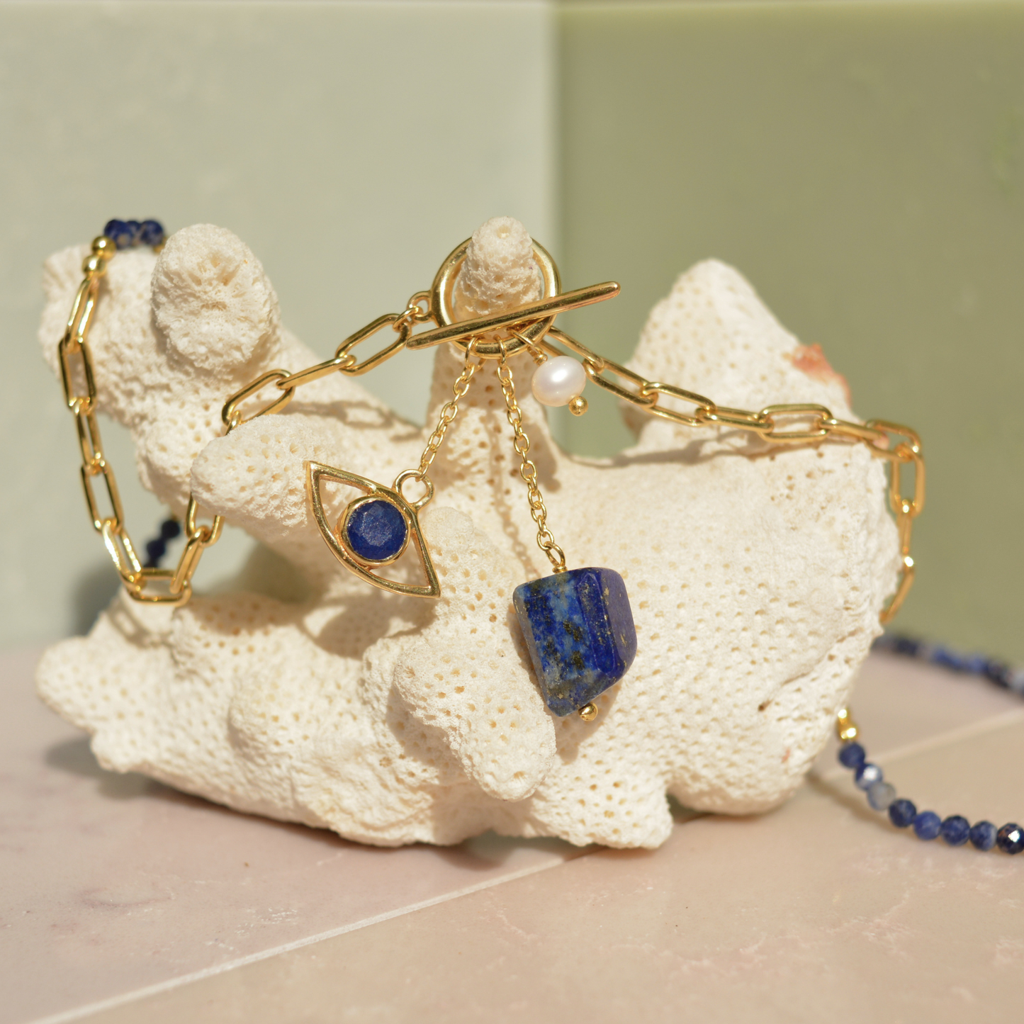 Close-up of the Meaningful Charm Necklace on a white coral prop, featuring the toggle clasp detail, natural lapis lazuli charms, and a delicate pearl accent.