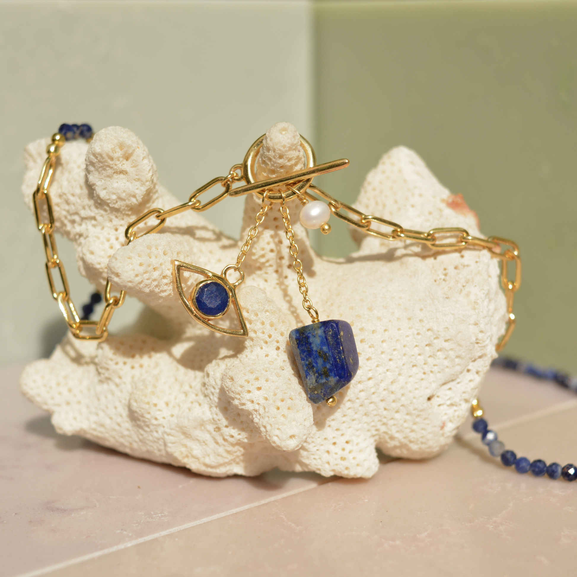 Close-up of the Meaningful Charm Necklace on a white coral prop, featuring the toggle clasp detail, natural lapis lazuli charms, and a delicate pearl accent.