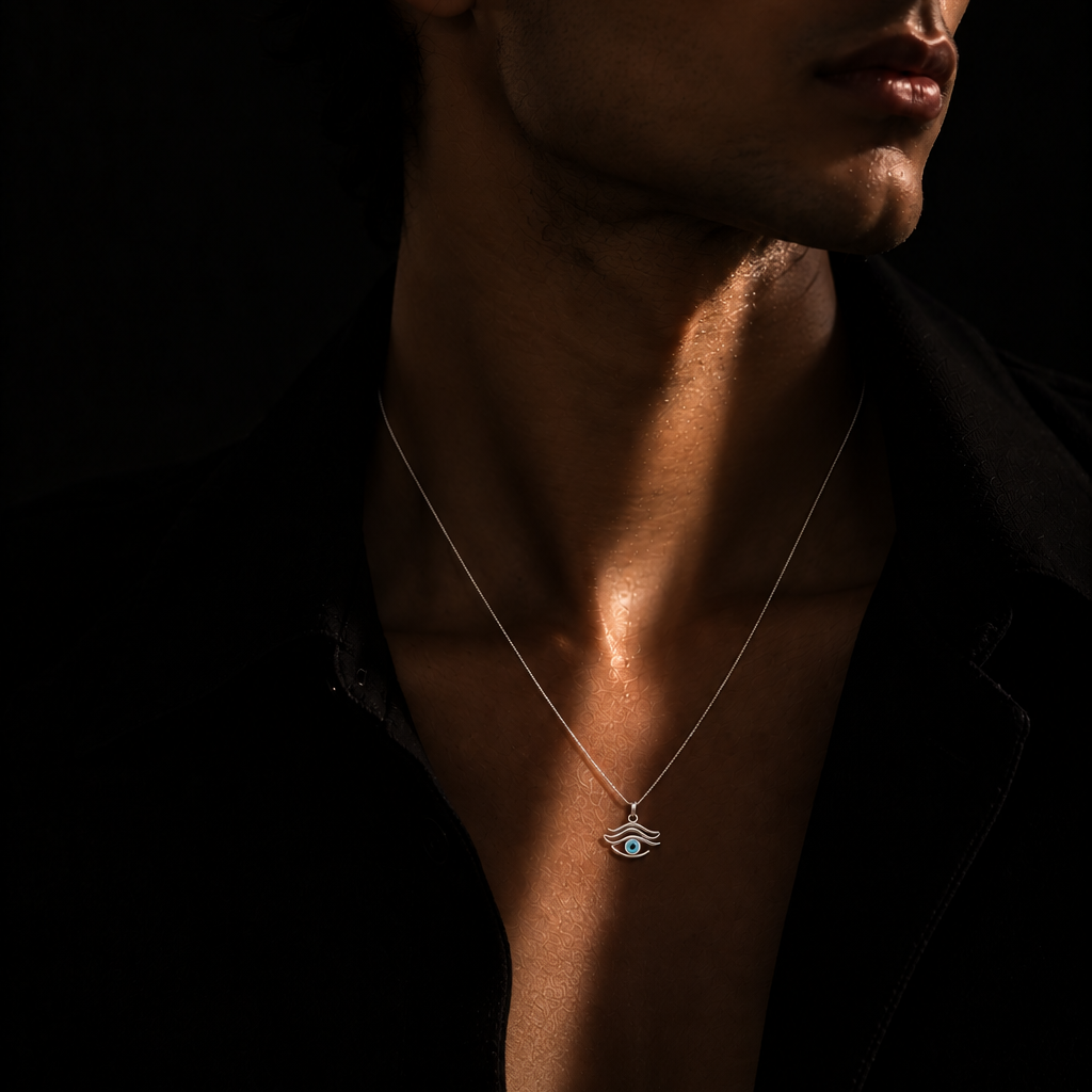 A man in a black shirt wearing a delicate silver chain necklace with a small evil eye pendant, captured in dramatic low-key lighting against a black background.