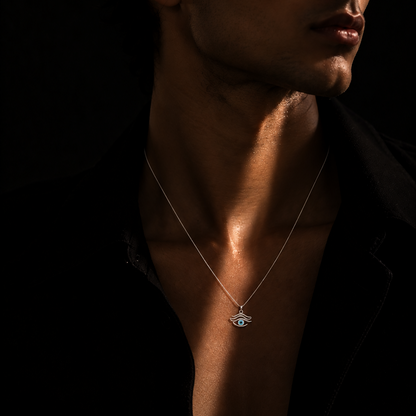 A man in a black shirt wearing a delicate silver chain necklace with a small evil eye pendant, captured in dramatic low-key lighting against a black background.