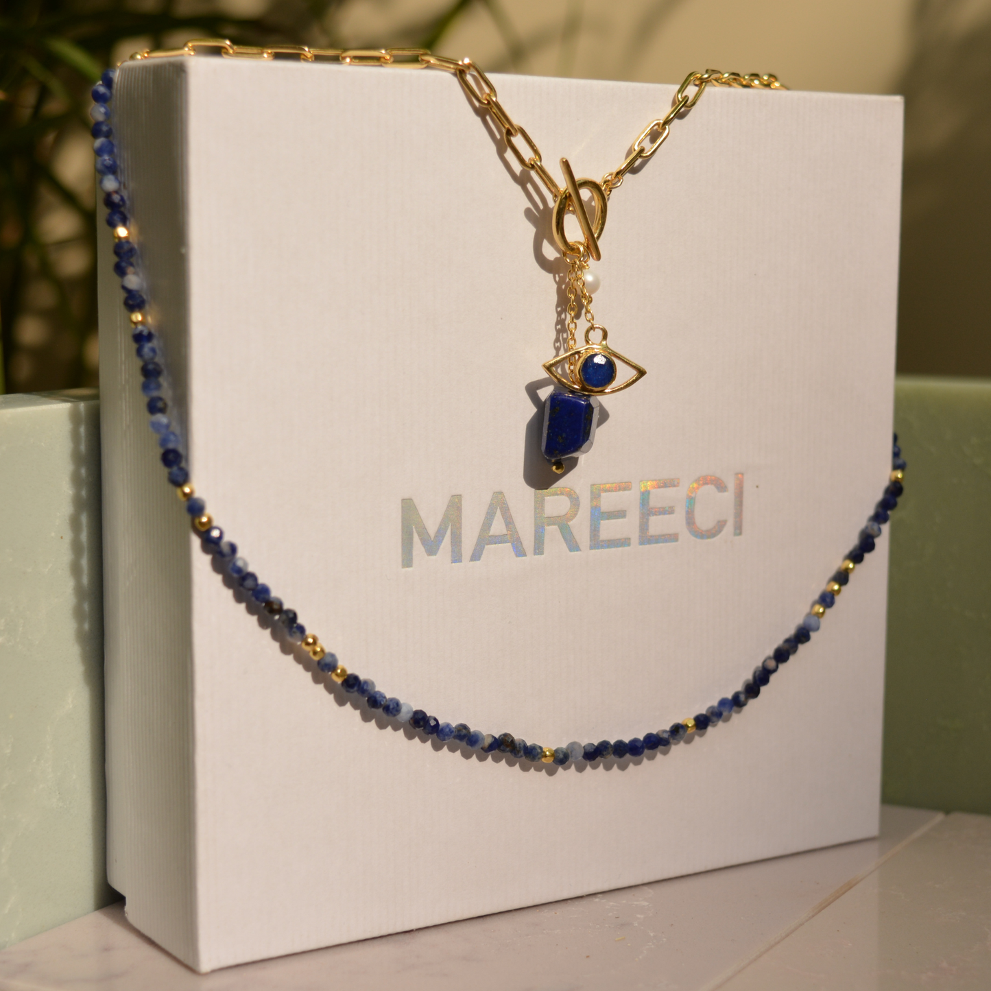 The Meaningful Charm Necklace in 18kt gold plating draped over a white MAREECI brand box, showcasing the blue lapis lazuli beads and evil eye talisman.