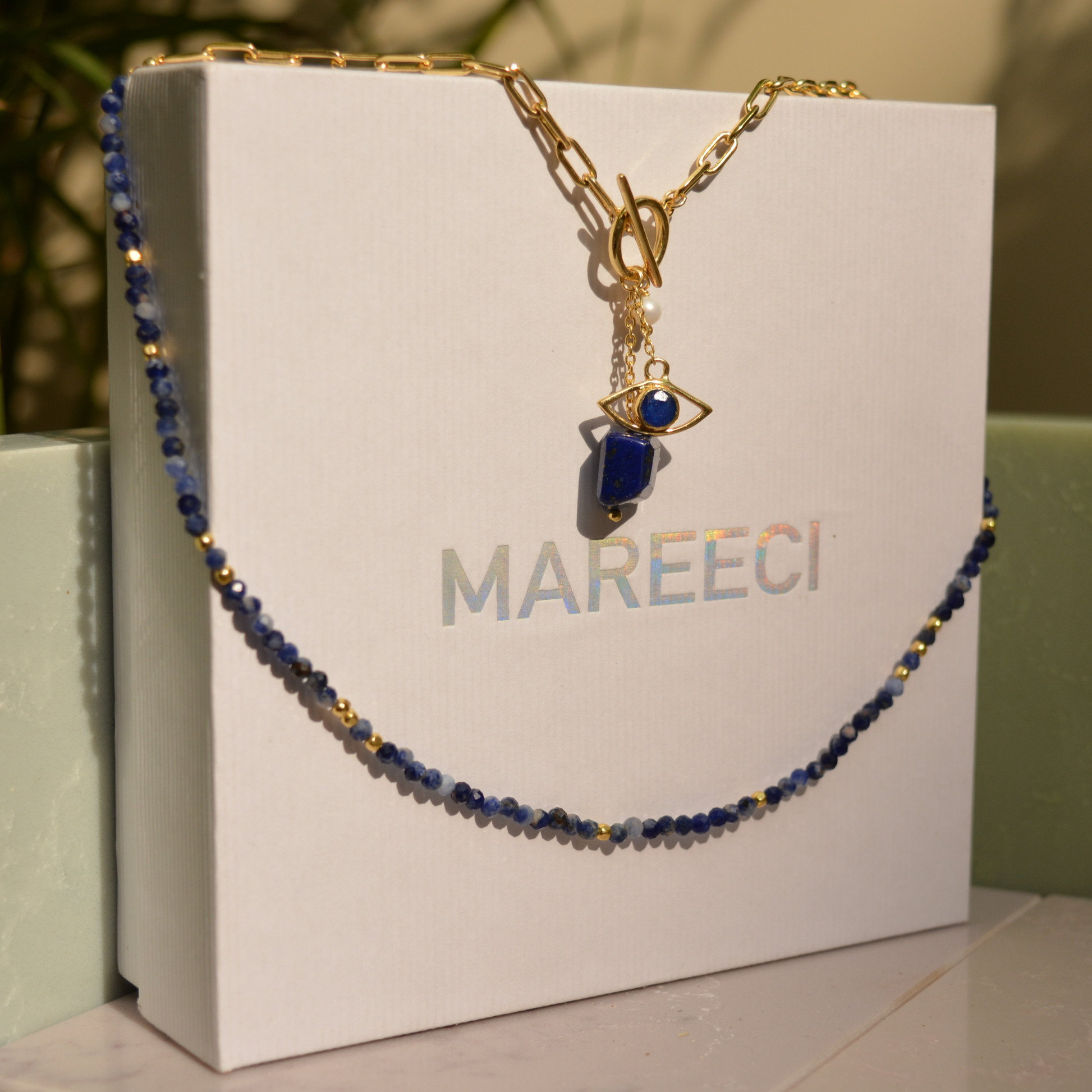The Meaningful Charm Necklace in 18kt gold plating draped over a white MAREECI brand box, showcasing the blue lapis lazuli beads and evil eye talisman.