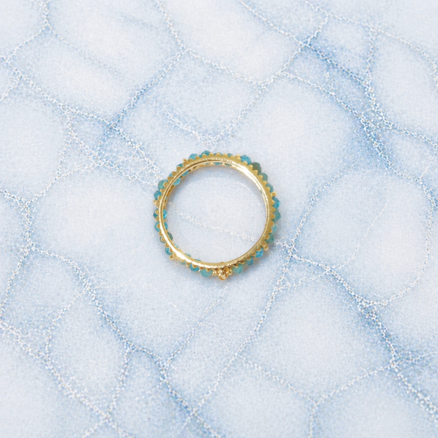 An overhead view of the Midnight Ball Ring, showcasing a continuous band of small blue-black jade beads set in 18kt gold plated 925 sterling silver on a blue marbled background.
