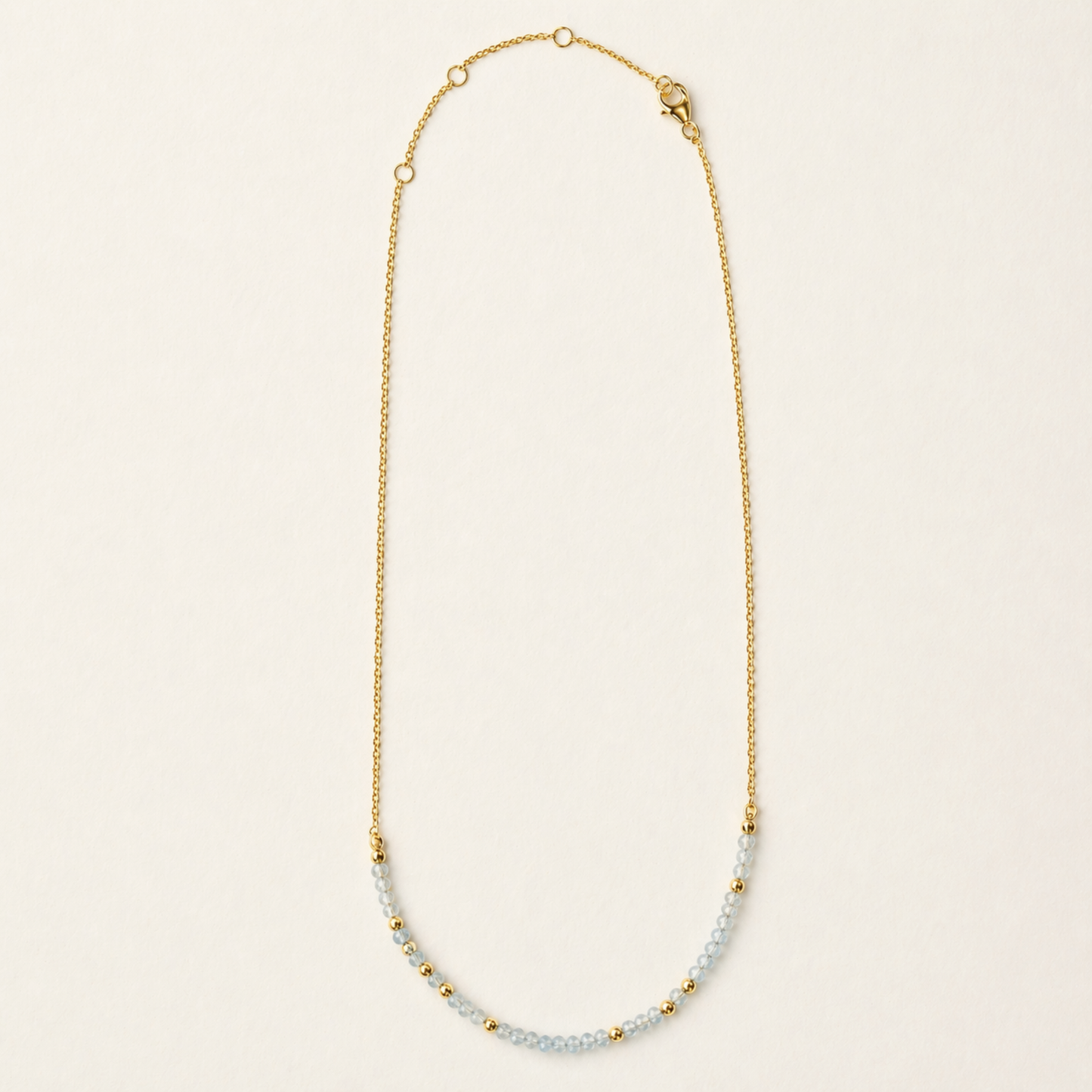Full studio shot of the Earthy Ease necklace on a cream background, showcasing the alternating pattern of natural round granite stones and tiny gold-plated beads on a delicate chain.