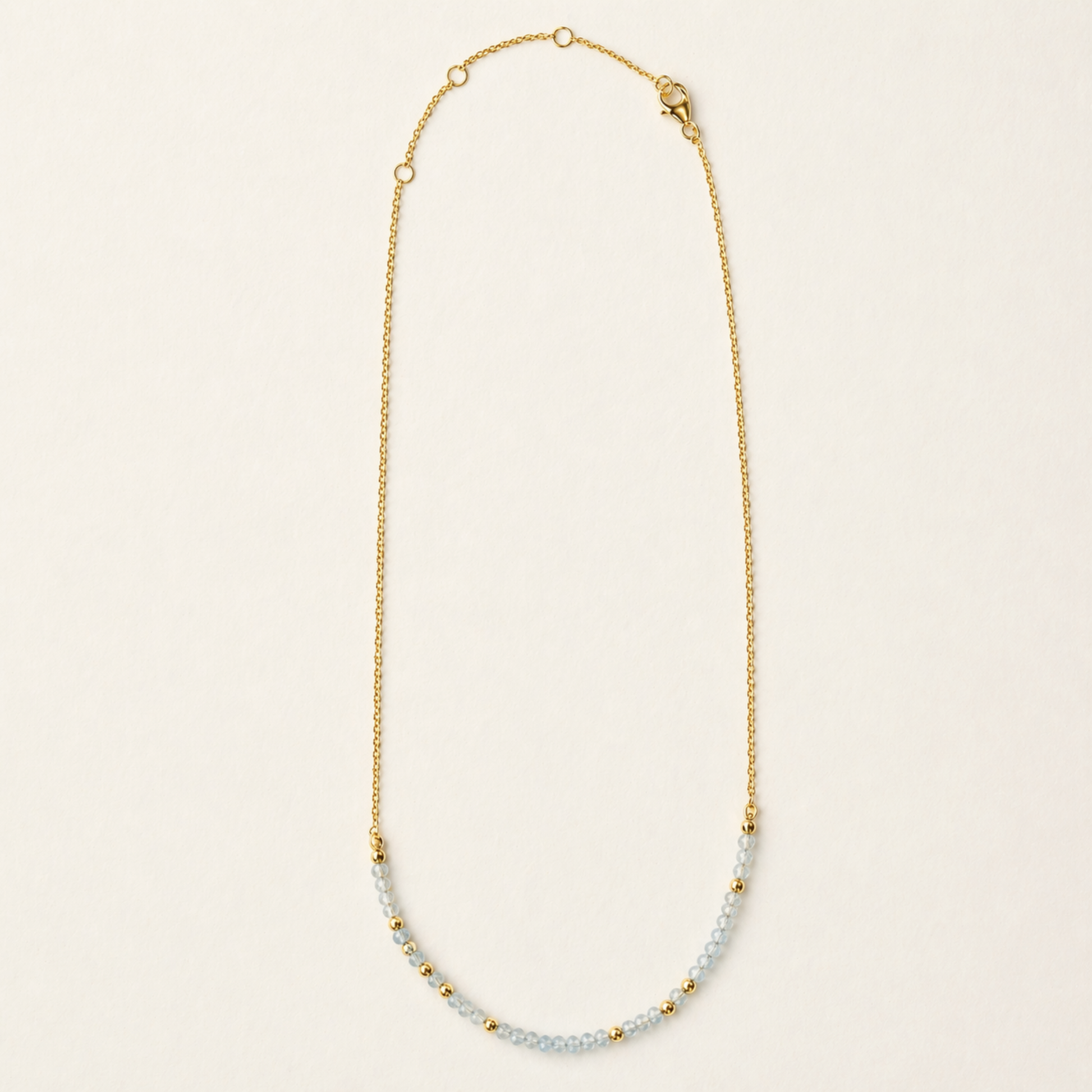 Full studio shot of the Earthy Ease necklace on a cream background, showcasing the alternating pattern of natural round granite stones and tiny gold-plated beads on a delicate chain.