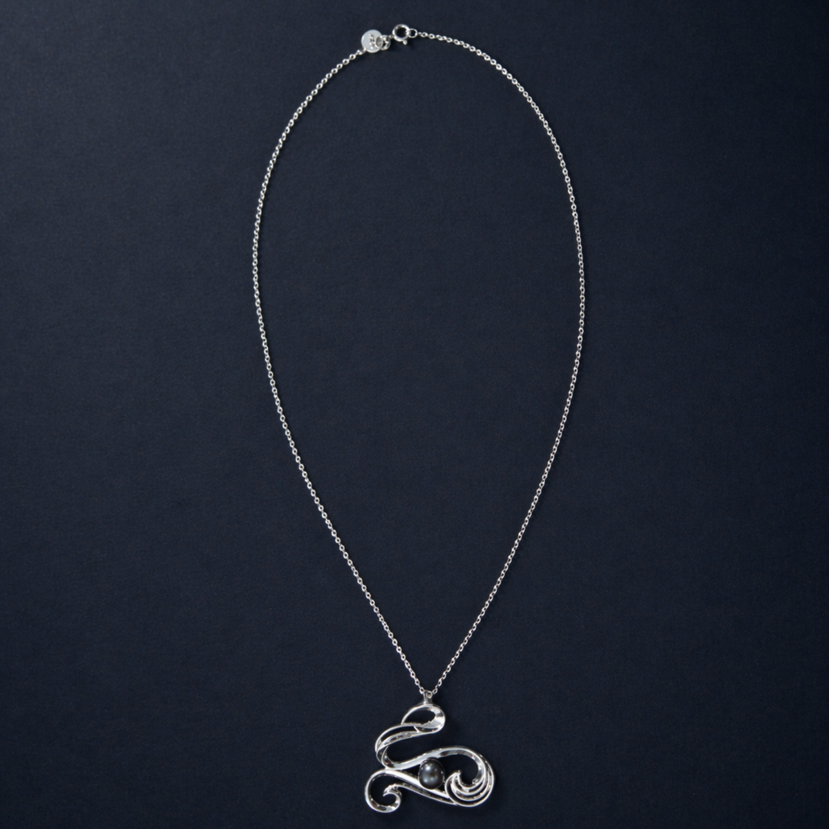 Full studio shot of the Graceful Flow Necklace on a dark navy background, showcasing the fluid, wavy silver pendant and the long, polished sterling silver chain.