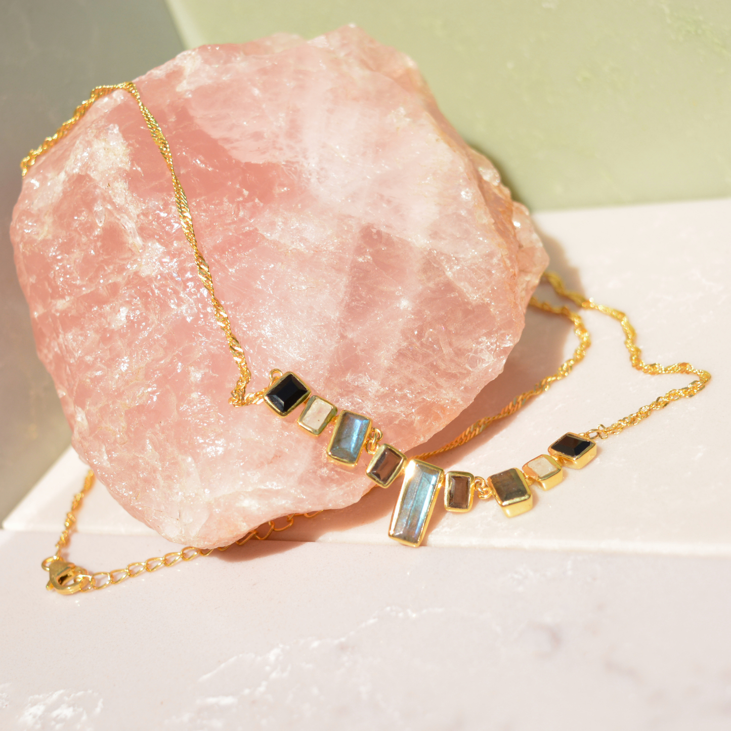 The A Story of Stones necklace draped over a large raw rose quartz crystal, highlighting the unique tones of the nine rectangular gemstone links and the twisted gold-plated chain.