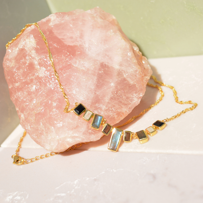 The A Story of Stones necklace draped over a large raw rose quartz crystal, highlighting the unique tones of the nine rectangular gemstone links and the twisted gold-plated chain.