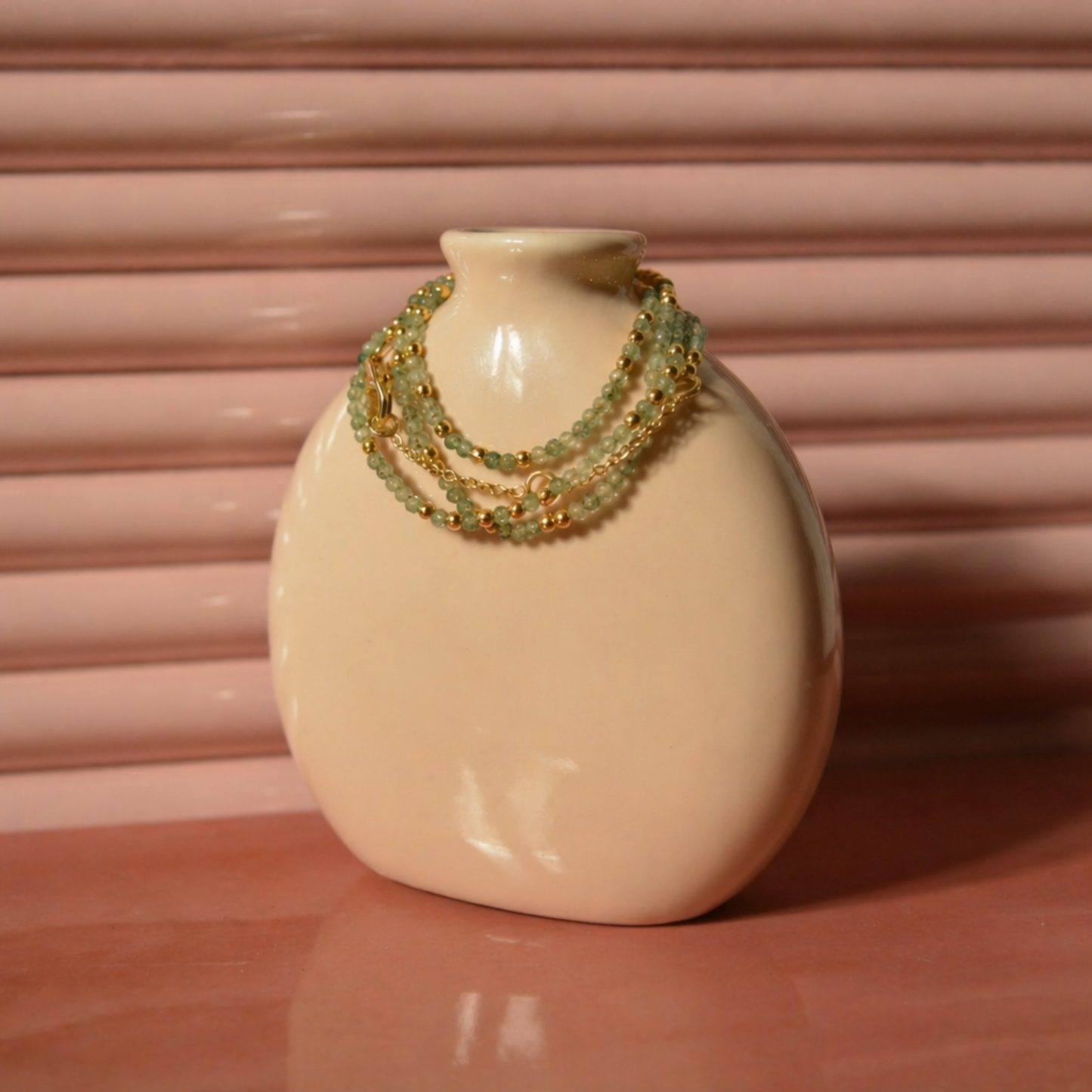 A handcrafted 18kt gold plated Nature’s Calm necklace featuring green beryl emerald and labradorite beads, wrapped around a minimal beige ceramic vase.