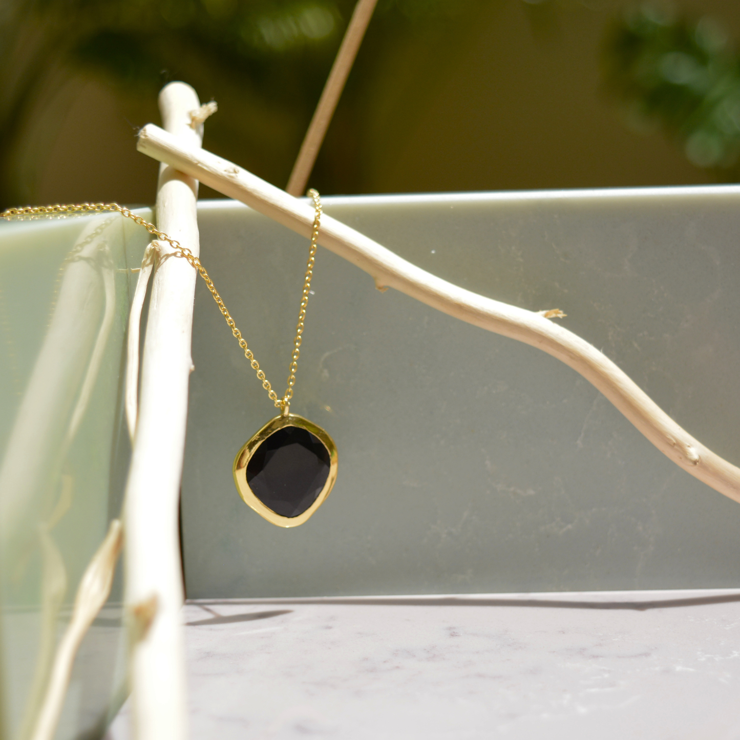 The Bold Calm black onyx necklace hanging from a light wooden branch, showcasing the modern organic shape and polished golden finish.