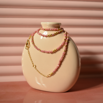 The Everyday Glow necklace wrapped around a minimal beige ceramic vase, highlighting the soft pink sparkle of the small ball-shaped beads and the warm 18kt gold finish.