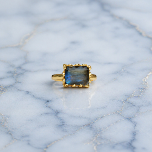 Frontal view of the Mystic Glow Ring featuring a thick rectangular labradorite stone with natural blue flashes, set in a polished gold plated minimal frame on marble.
