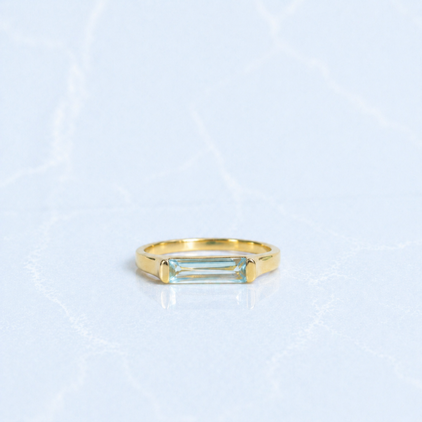A handcrafted 18kt gold plated Sky Glow Ring featuring a rectangular blue topaz stone with a soft sky-blue hue, set in a polished minimal 925 sterling silver band on a light marbled background.